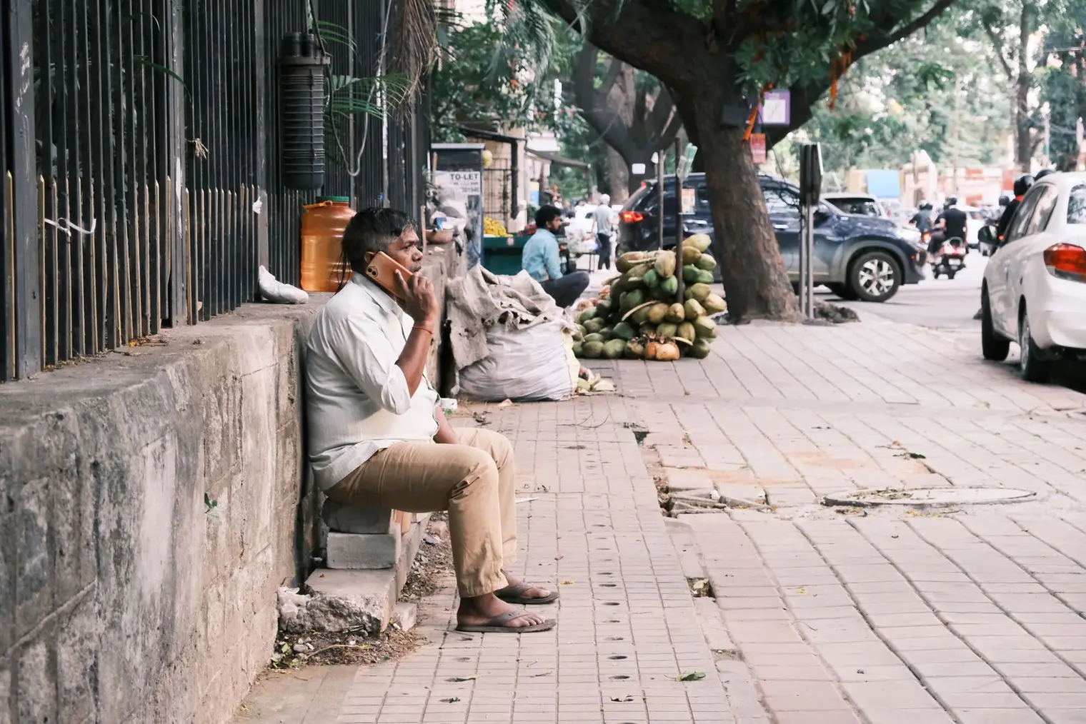 A man sits on a kerb on a Bengaluru pavement, talking on his phone, with a coconut seller behind him