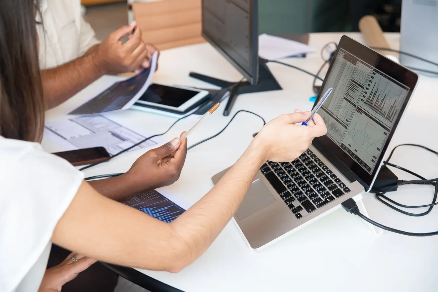 Two colleagues pointing at charts on a laptop while reviewing printed reports at a desk