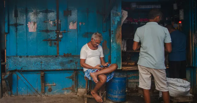 An older man sits on a stool in front of a blue wooden shopfront, looking at his phone